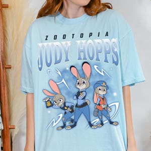 May include: Pale blue t-shirt with the text "ZOOTOPIA JUDY HOPPS" and a graphic of three cartoon bunnies in police uniforms. The bunnies are in shades of grey, blue, and orange. The shirt has a crew neck.