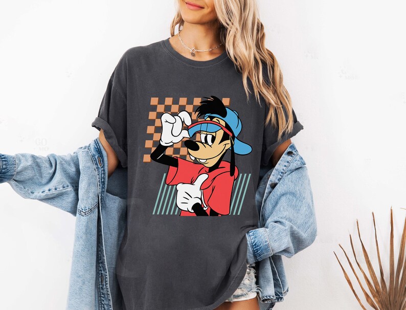 May include: A dark grey t-shirt featuring a cartoon image of Goofy wearing a blue baseball cap and red shirt. The image is set against a checkered background with green and blue stripes.