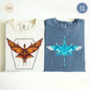 May include: Two t-shirts, one ivory and one blue jean, each with a unique bird graphic inside a hexagon. The ivory shirt features an orange and yellow bird design, while the blue jean shirt displays a blue and white bird.