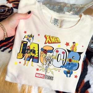 May include: Cream-colored sweatshirt featuring the word "JACOB" in large, colorful letters with X-Men characters and the "X-MEN" logo. The Marvel Studios logo is at the bottom. The sweatshirt is held by a person.