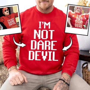 Peut inclure: Un sweat-shirt rouge avec le texte "I'M NOT DARE DEVIL" en lettres blanches. Deux images encadrées d'un personnage de bande dessinée sont de chaque côté du sweat-shirt. Le sweat-shirt est à manches longues.
