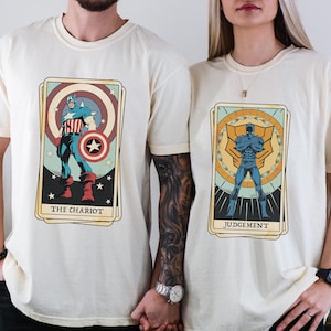 May include: Two cream-colored t-shirts with graphic designs of tarot cards featuring superheroes. The left shirt features Captain America as "The Chariot" and the right shirt features a blue superhero as "Judgement".