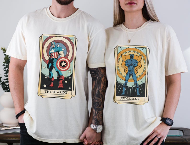 May include: Two cream-colored t-shirts with graphic designs of tarot cards featuring superheroes. The left shirt features Captain America as "The Chariot" and the right shirt features a blue superhero as "Judgement".