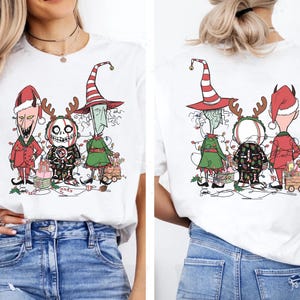 K&ouml;nnte beinhalten: Wei&szlig;es T-Shirt mit einer Cartoon-Illustration von drei Figuren in Weihnachtsoutfits. Die Figuren tragen einen roten Weihnachtsmannhut, einen Hexenhut und ein Rentier-Geweih-Stirnband. Das Design umfasst Lichterketten und Geschenkboxen.