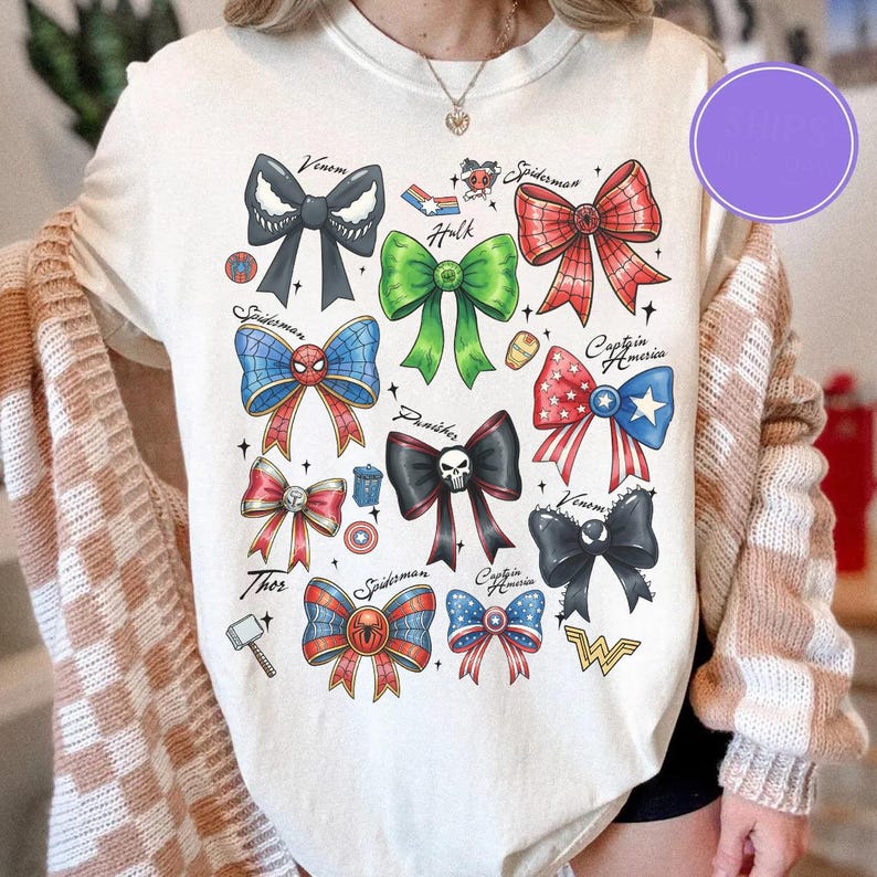 May include: Cream-colored t-shirt featuring colorful bow designs inspired by Marvel characters like Spider-Man, Captain America, and Venom. Each bow is uniquely colored and styled, with character names above. A fun and creative graphic tee.