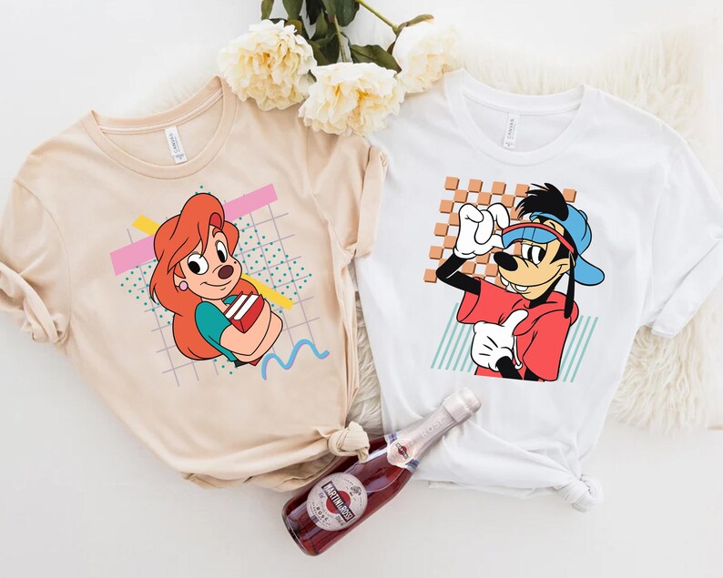 May include: Two t-shirts featuring cartoon characters. The first t-shirt is beige with a cartoon of a female dog with red hair holding books. The second t-shirt is white with a cartoon of a dog wearing a blue hat and red shirt. Both t-shirts have a geometric pattern in the background.