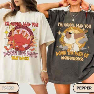 Könnte beinhalten: Zwei T-Shirts, eines in Elfenbein und eines in Pfeffer, mit grafischen Designs. Das elfenbeinfarbene Shirt zeigt eine rote Figur mit einem Dreizack und dem Text "I'M GONNA LEAD YOU DOWN THE PATH THAT ROCKS." Das pfefferfarbene Shirt hat eine weiße Figur und den Text "I'M GONNA LEAD YOU DOWN THE PATH OF RIGHTEOUSNESS."
