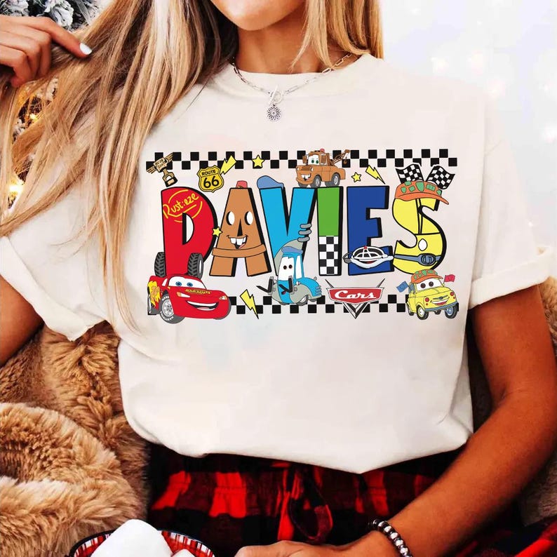 May include: Cream-colored t-shirt featuring the name "DAVIES" in colorful, cartoon-style lettering with images of characters from the movie "Cars." The design includes a Route 66 sign and checkered flag accents.