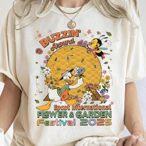 Puede incluir: Camiseta color crema con un diseño de dibujos animados con el Pato Donald, abejas y flores. El texto dice "BUZZIN' Around" y "Epcot International Flower & Garden Festival 2025". El diseño incluye un panal y elementos florales.