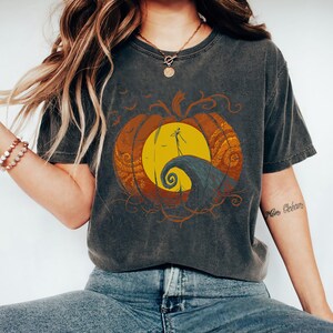 Pode incluir: Uma camiseta cinza escura com um gr&aacute;fico de estilo vintage de uma ab&oacute;bora com uma cara de jack-o'-lantern. A ab&oacute;bora &eacute; cercada por um design laranja e amarelo em espiral. O texto "This is Halloween" est&aacute; impresso na camiseta.