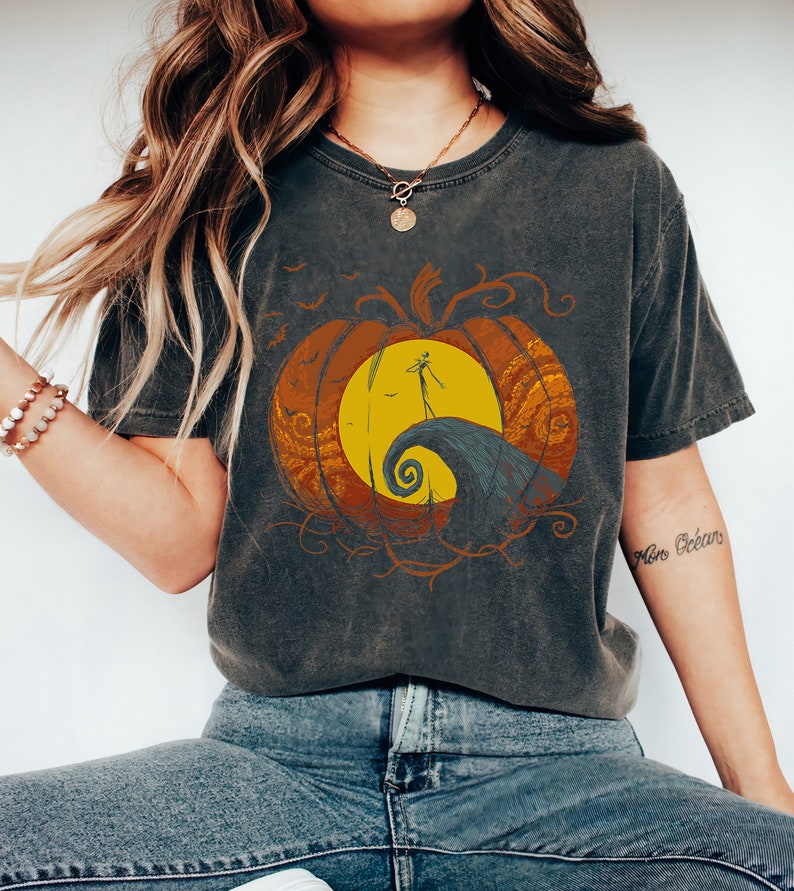 Pode incluir: Uma camiseta cinza escura com um gr&aacute;fico de estilo vintage de uma ab&oacute;bora com uma cara de jack-o'-lantern. A ab&oacute;bora &eacute; cercada por um design laranja e amarelo em espiral. O texto "This is Halloween" est&aacute; impresso na camiseta.