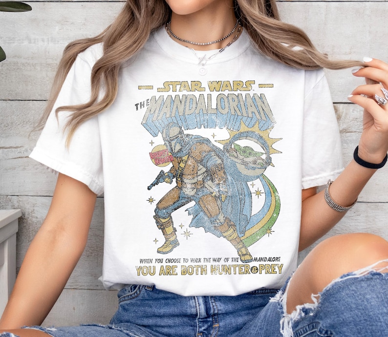 K&ouml;nnte beinhalten: Wei&szlig;es T-Shirt mit einem Vintage-Comic-Grafik eines Mandalorianer-Charakters aus Star Wars. Der Text lautet "STAR WARS - THE MANDALORIAN" und "WHEN YOU CHOOSE TO WALK THE WAY OF THE MANDALORE YOU ARE BOTH HUNTER PREY".