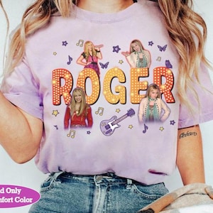 May include: A light purple t-shirt featuring the word "ROGER" in large, illuminated letters. The shirt has images of a singer and musical notes, stars, and a guitar. The shirt is made of a soft, comfortable material.