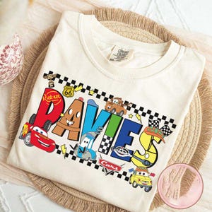 May include: Cream-colored t-shirt featuring the name "Davies" in colorful, cartoon-style lettering with images of characters from the movie Cars. The design includes Lightning McQueen, Mater, and other characters, along with racing flags and Route 66 signage.