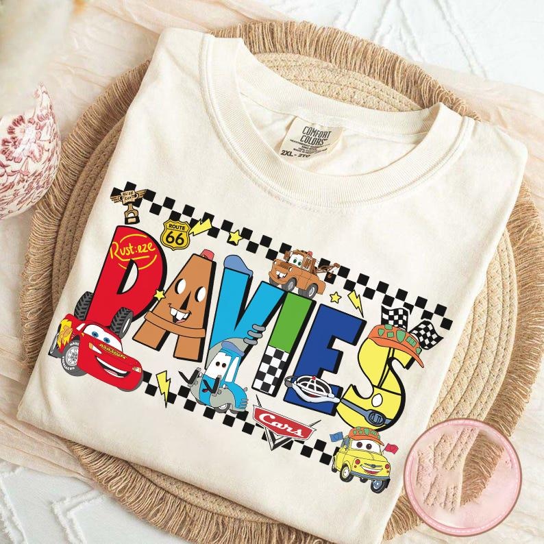 May include: Cream-colored t-shirt featuring the name "Davies" in colorful, cartoon-style lettering with images of characters from the movie Cars. The design includes Lightning McQueen, Mater, and other characters, along with racing flags and Route 66 signage.