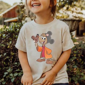 Puede incluir: Camiseta beige con una ilustraci&oacute;n de dibujos animados de un personaje de rat&oacute;n sosteniendo un helado con forma de Mickey Mouse. El rat&oacute;n lleva un sombrero negro y un abrigo naranja. La camiseta es de tela suave de color neutro.
