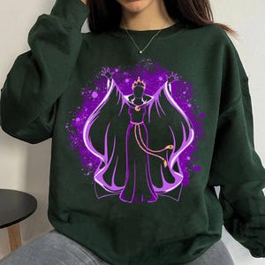 May include: A dark green sweatshirt featuring a purple and black silhouette of a woman with a crown and a long flowing cape. The woman is holding her arms out to the sides. The design is in a watercolour style.