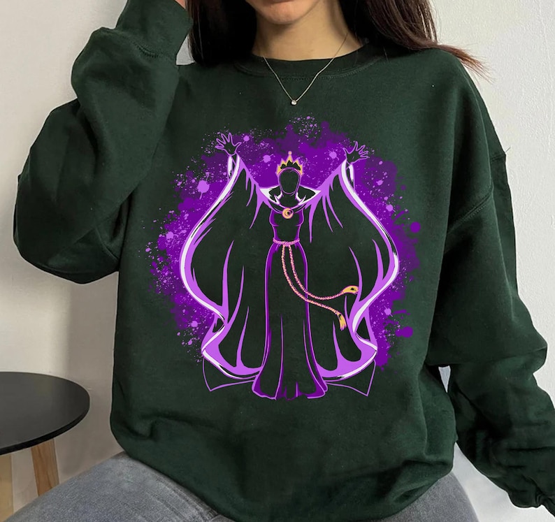 May include: A dark green sweatshirt featuring a purple and black silhouette of a woman with a crown and a long flowing cape. The woman is holding her arms out to the sides. The design is in a watercolour style.