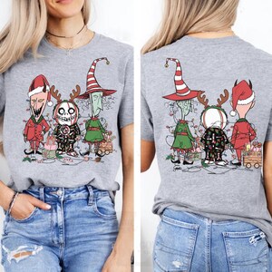 K&ouml;nnte beinhalten: Hellgraues T-Shirt mit einer Grafik von drei Cartoon-Charakteren in Weihnachtsoutfits. Vorder- und R&uuml;ckseite des Shirts sind abgebildet. Die Charaktere sind mit Weihnachtsbeleuchtung geschm&uuml;ckt und stehen vor verpackten Geschenken.
