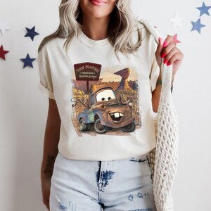 Może przedstawiać: Biała koszulka z grafiką Tow Matera z filmu Auta. Grafika przedstawia Tow Matera w pustynnym otoczeniu z tabliczką z napisem "Tow Mater - Radiator Springs - Tow Truck Service".