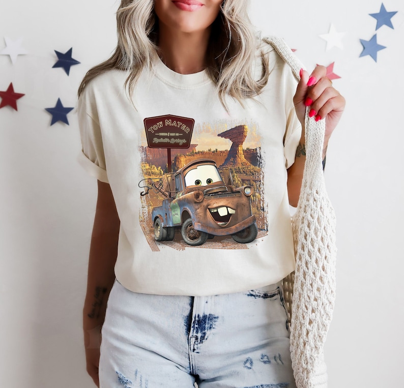Może przedstawiać: Biała koszulka z grafiką Tow Matera z filmu Auta. Grafika przedstawia Tow Matera w pustynnym otoczeniu z tabliczką z napisem "Tow Mater - Radiator Springs - Tow Truck Service".