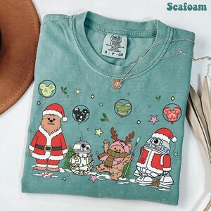 Puede incluir: Camiseta verde mar con un gráfico festivo que presenta personajes de Star Wars vestidos para Navidad. Chewbacca con traje de Papá Noel, BB-8, un Ewok y R2-D2 están representados. Globos de Mickey Mouse y adornos navideños completan el diseño.
