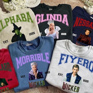 May include: A collection of crewneck sweatshirts in various colors, each featuring a character from the musical "Wicked." The sweatshirts display character names in collegiate lettering, along with images of the characters and the word "Wicked" at the bottom.