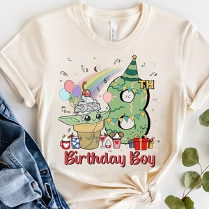 May include: A beige t-shirt with a green and white cartoon character holding a green ice cream cone. The character is standing next to a green number eight with a party hat. The text "Birthday Boy" is printed in red below the characters.