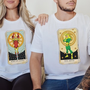 May include: Two white t-shirts with a tarot card design. The left shirt features a character in a red and gold suit with the text "King of Coins". The right shirt features a character in a green suit with the text "The Devil".