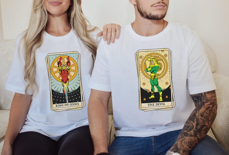 May include: Two white t-shirts with a tarot card design. The left shirt features a character in a red and gold suit with the text "King of Coins". The right shirt features a character in a green suit with the text "The Devil".