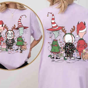 K&ouml;nnte beinhalten: Helllila T-Shirt mit einer Cartoon-Illustration von drei festlichen Charakteren. Sie sind mit Weihnachtsbeleuchtung geschm&uuml;ckt und stehen in der N&auml;he eines kleinen Wagens voller Geschenke. Das Design ist auf der Vorder- und R&uuml;ckseite des T-Shirts aufgedruckt.