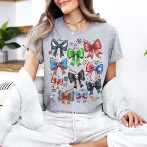 May include: A gray t-shirt featuring colorful bow designs with superhero themes. The bows are in various colors and patterns, with text labels. The shirt is made of a soft material.