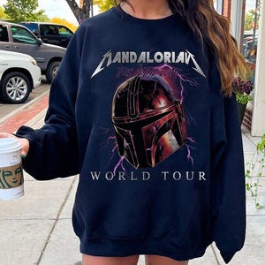 May include: Navy blue sweatshirt featuring a graphic of a Mandalorian helmet with lightning bolts and the text "MANDALORIAN THIS IS THE WAY WORLD TOUR" in silver and white. The sweatshirt is a crew neck style.