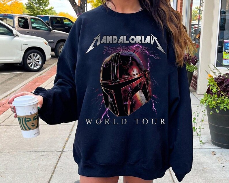 May include: Navy blue sweatshirt featuring a graphic of a Mandalorian helmet with lightning bolts and the text "MANDALORIAN THIS IS THE WAY WORLD TOUR" in silver and white. The sweatshirt is a crew neck style.