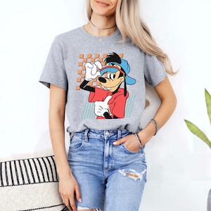 May include: A grey t-shirt featuring a cartoon image of Goofy wearing a blue baseball cap and red shirt. The image is set against a light blue and white checkered background.