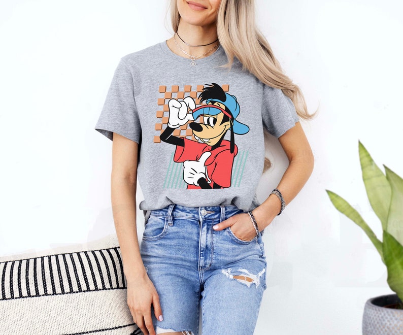 May include: A grey t-shirt featuring a cartoon image of Goofy wearing a blue baseball cap and red shirt. The image is set against a light blue and white checkered background.