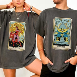 May include: Two gray t-shirts with a tarot card design. The left shirt features a red and yellow illustration of "The Witch" with the text "The Witch". The right shirt features a yellow and blue illustration of "The Emperor" with the text "Emperor".