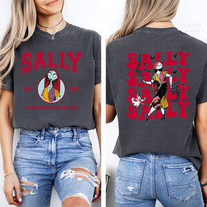 K&ouml;nnte beinhalten: Ein dunkelgraues T-Shirt mit einem roten Grafikdruck von Sally aus Nightmare Before Christmas. Die Vorderseite des Shirts tr&auml;gt den Schriftzug "Sally Est. 1993 The Nightmare Before Christmas". Die R&uuml;ckseite des Shirts zeigt das Wort "Sally" mehrmals in einer roten, stilisierten Schrift.