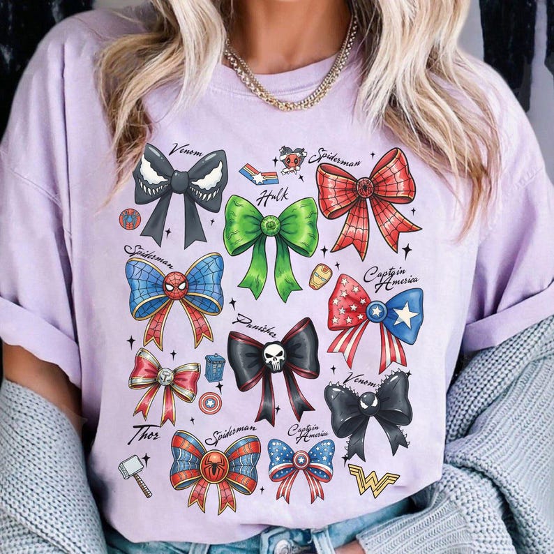 May include: A light purple t-shirt featuring a collection of colorful bow designs inspired by Marvel characters, including Spider-Man, Hulk, and Captain America. Each bow is uniquely colored and styled, with character names written above.