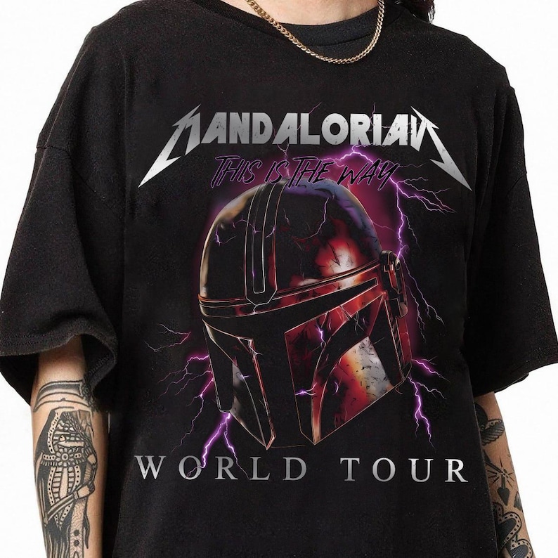 May include: Black t-shirt featuring a graphic of a Mandalorian helmet with purple lightning and the text "MANDALORIAN" and "THIS IS THE WAY". Below the helmet is the text "WORLD TOUR" in white.