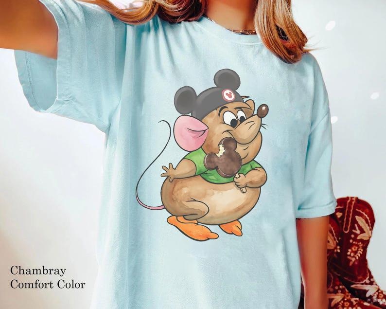 Puede incluir: Camiseta azul claro con un rat&oacute;n de dibujos animados que lleva un sombrero de Mickey Mouse y come un helado con forma de Mickey Mouse. El rat&oacute;n es marr&oacute;n, con una oreja rosa y pies naranjas. La camiseta tiene la inscripci&oacute;n "Chambray Comfort Color".