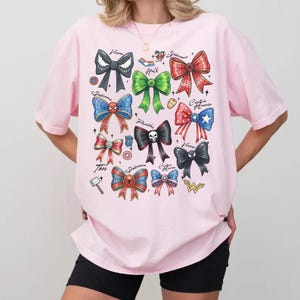 May include: A light pink t-shirt featuring a colorful graphic of superhero-themed bows. The bows are in various colors, including red, blue, green, and black, with superhero logos and names. The shirt is a casual, everyday wear item.