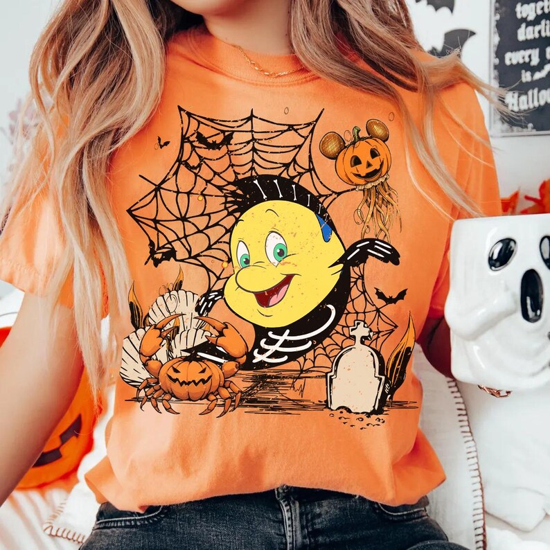 May include: Orange t-shirt featuring a Halloween-themed graphic. The design includes a yellow cartoon character with a skeleton outline, spiderwebs, pumpkins, and a crab. A pumpkin with Mickey Mouse ears is also present.