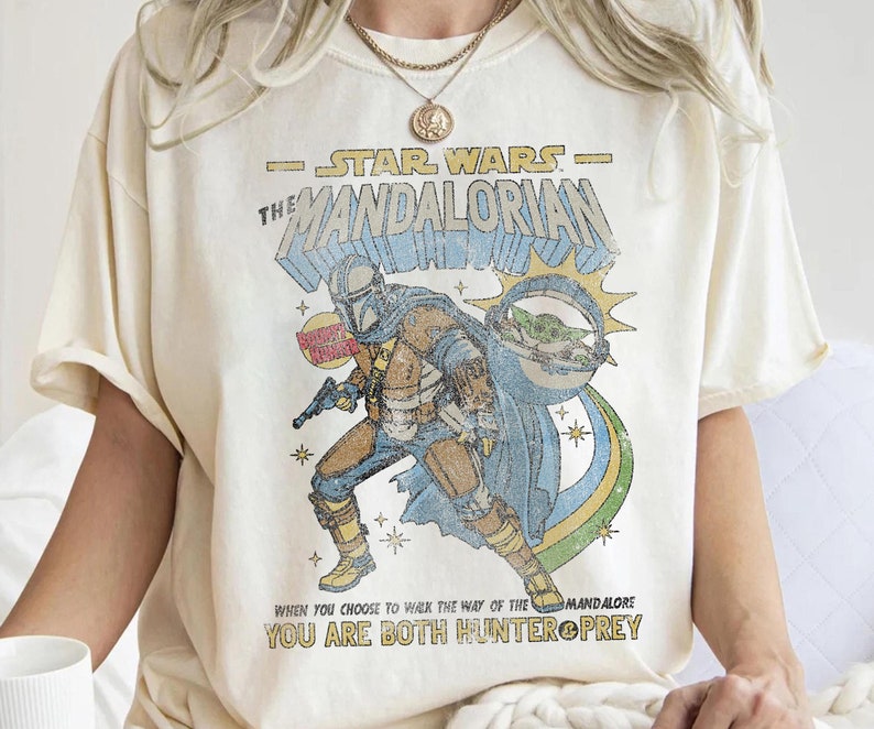 K&ouml;nnte beinhalten: Ein wei&szlig;es T-Shirt mit einer Grafik im Vintage-Stil eines Charakters aus Star Wars: The Mandalorian. Die Grafik zeigt den Mandalorianer, Grogu und den Text "STAR WARS - THE MANDALORIAN" und "WHEN YOU CHOOSE TO WALK THE WAY OF THE MANDALORE YOU ARE BOTH HUNTER & PREY".