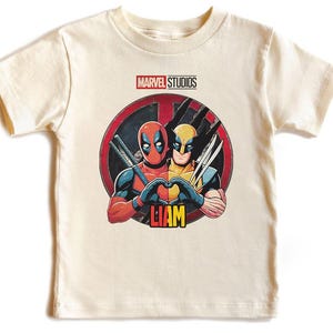 May include: Cream-colored t-shirt featuring a graphic of Deadpool and Wolverine forming a heart shape. The Marvel Studios logo is at the top, with the name "LIAM" in yellow at the bottom. The design is set against a red and black circular background.