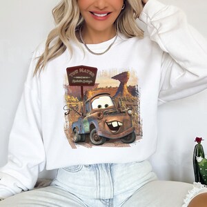 Może przedstawiać: Biała bluza z nadrukiem Tow Matera z filmu Auta. Grafika przedstawia Tow Matera jadącego po drodze gruntowej ze znakiem, na kt&oacute;rym widnieje napis "Tow Mater - Witamy w Radiator Springs".