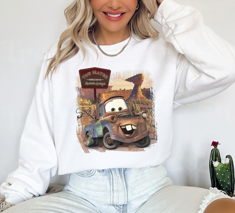 Może przedstawiać: Biała bluza z nadrukiem Tow Matera z filmu Auta. Grafika przedstawia Tow Matera jadącego po drodze gruntowej ze znakiem, na kt&oacute;rym widnieje napis "Tow Mater - Witamy w Radiator Springs".