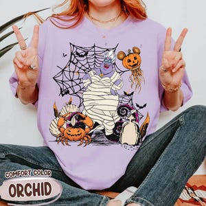 May include: A lilac Comfort Color t-shirt featuring a Halloween-themed graphic. The design includes a cartoon Ursula character wrapped in bandages, surrounded by pumpkins, spiderwebs, and bats. The word "ORCHID" is printed on the bottom.