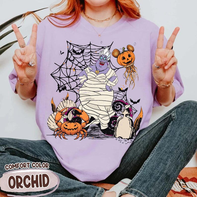 May include: A lilac Comfort Color t-shirt featuring a Halloween-themed graphic. The design includes a cartoon Ursula character wrapped in bandages, surrounded by pumpkins, spiderwebs, and bats. The word "ORCHID" is printed on the bottom.
