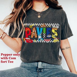 May include: Dark gray t-shirt featuring the name "DAVIES" in colorful, cartoon-style lettering with car graphics. The shirt has a distressed look and includes the text "Pepper only with Comfort Tee".